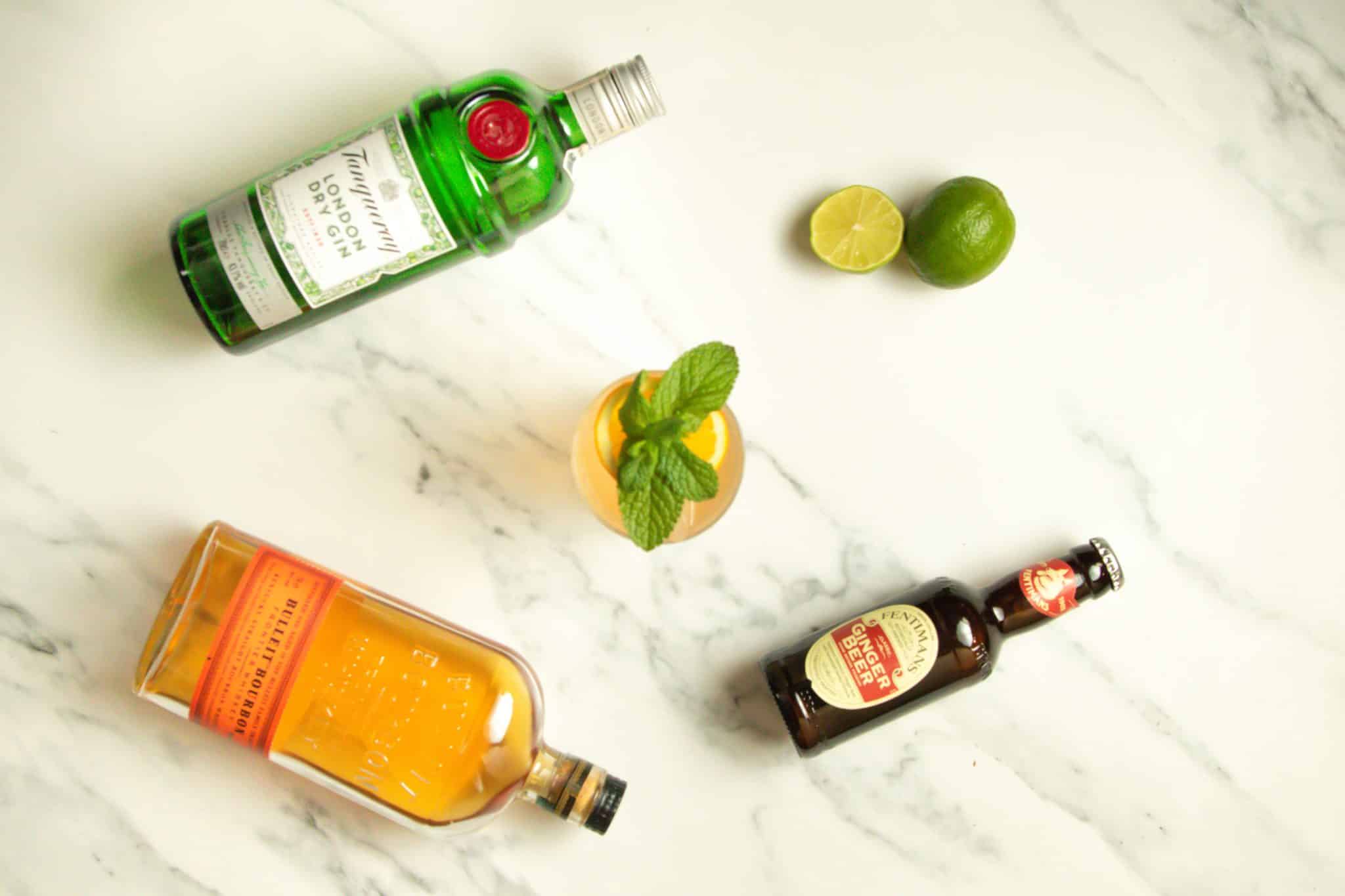 A Suffering Bastard cocktail, shot from above, on a white marmol table with a Gin bottle, a Bourbon bottle, a ginger beer bottle and two limes around.