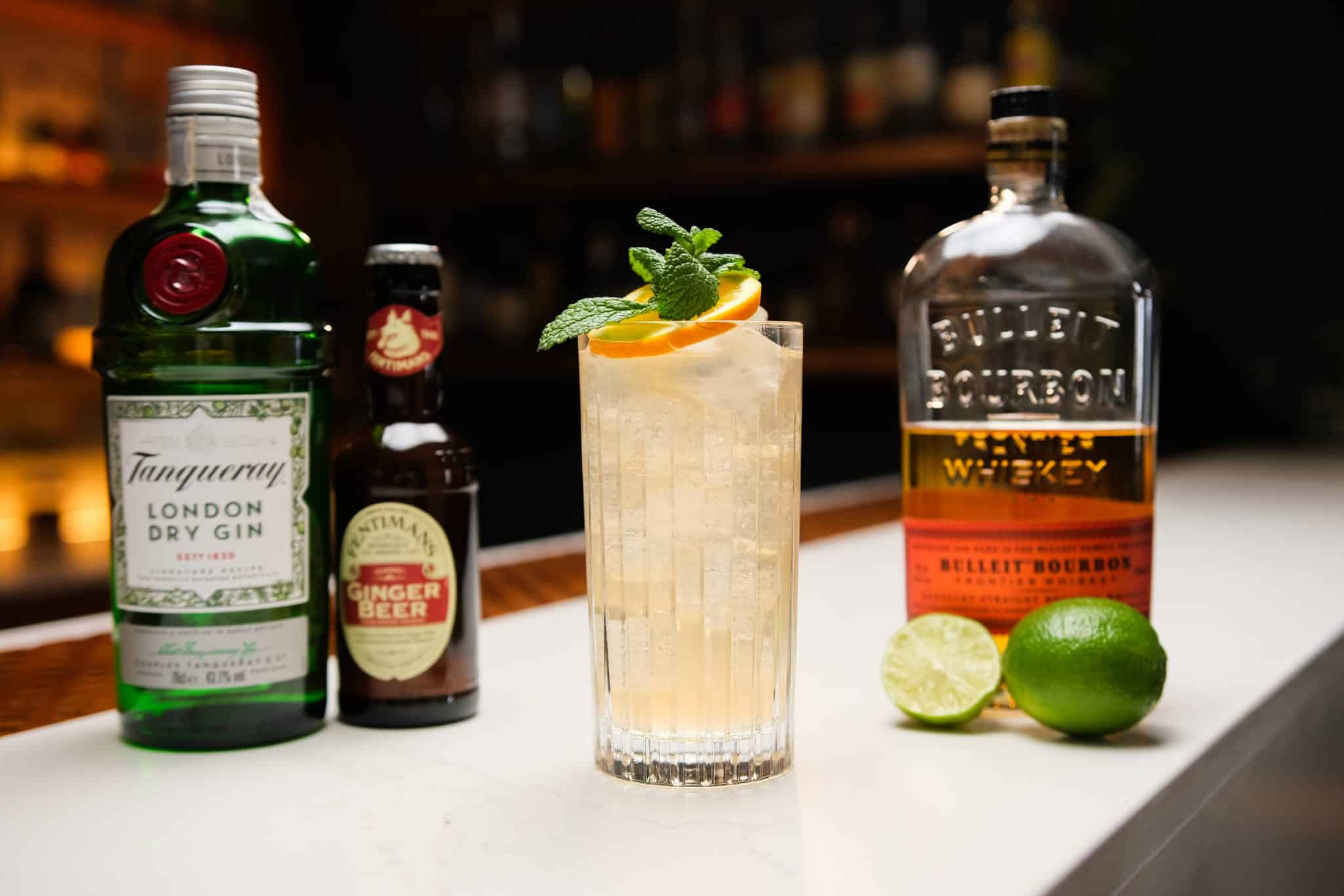 Bourbon, Gin, Ginger beer and two limes laid out on a white bar table.