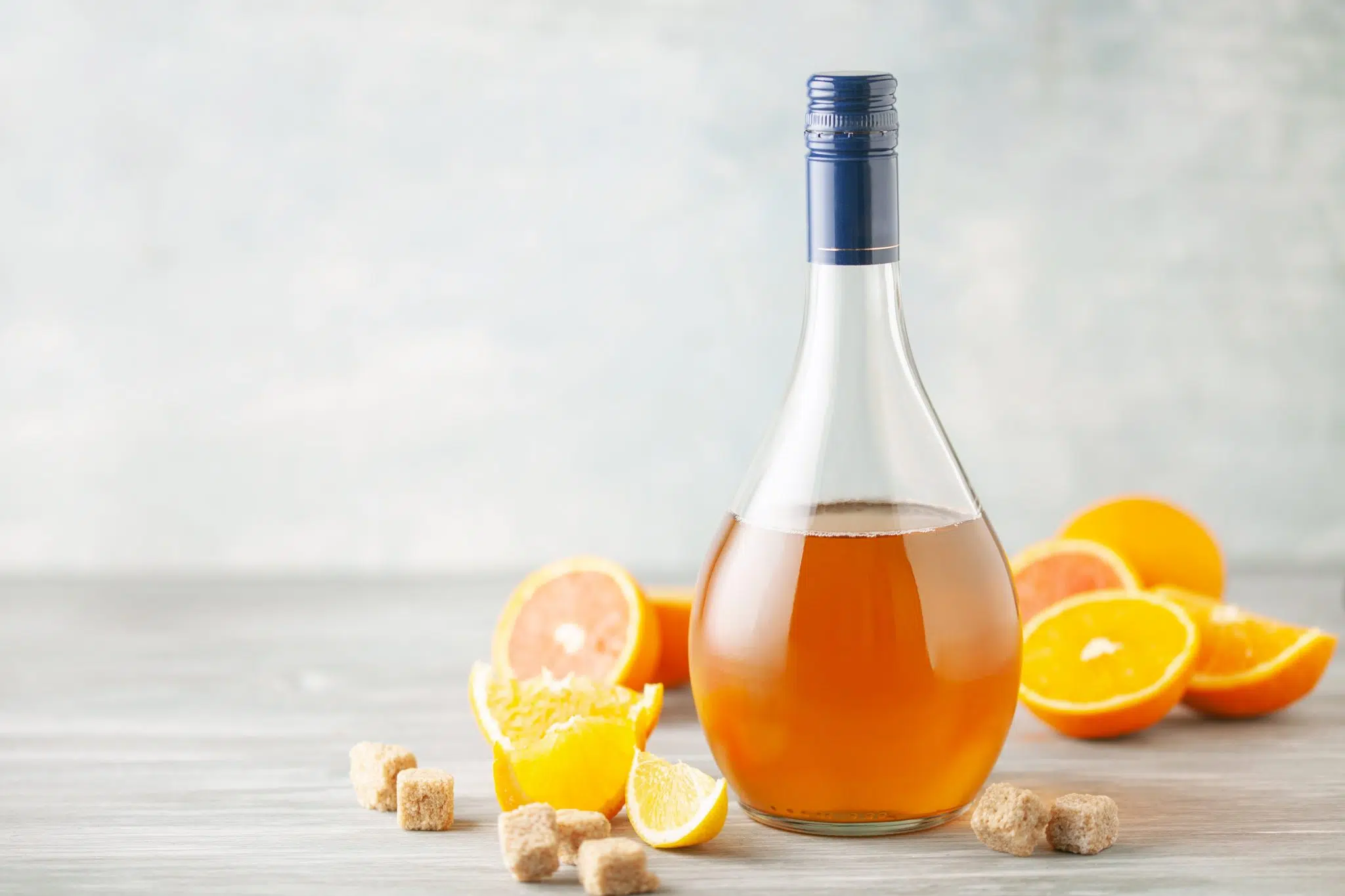 Shot of a homemade orange liqueur bottle on a grey wooden surface surrounded by orange wedges and slices and five brown sugar cubes