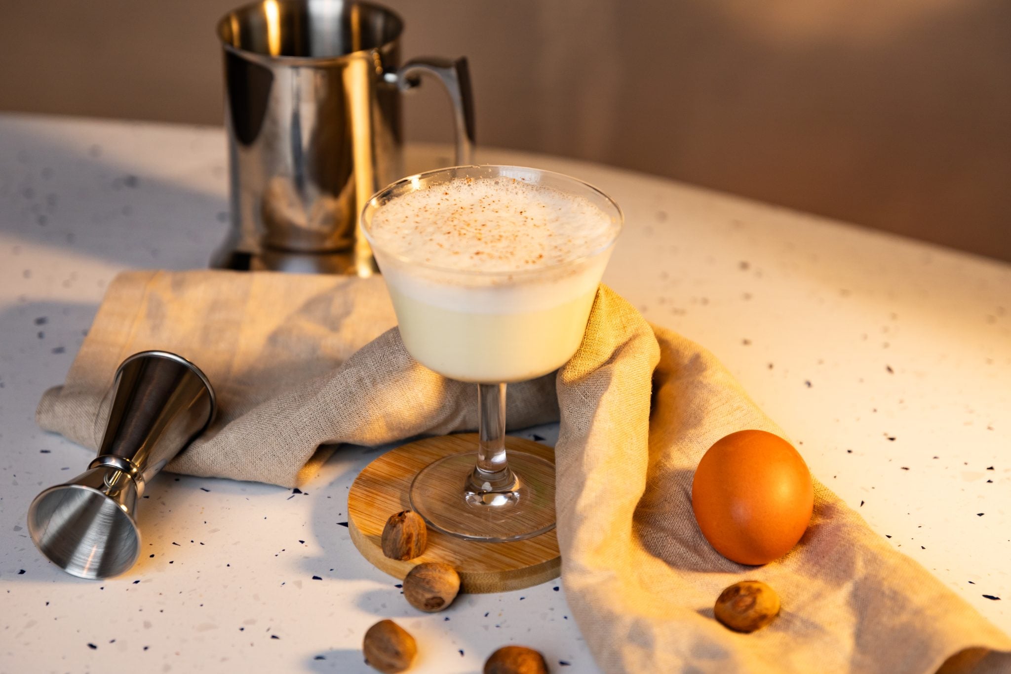 A side shot of a Suissesse cocktail in a coupe glass on a wooden coaster placed on a white surface surrounded by a jigger, many nutmegs, an egg, a beige cloth and a stainless steel jug