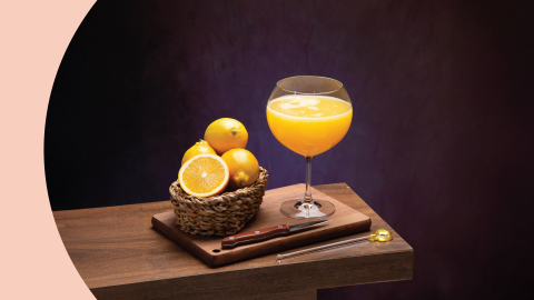 A shot of a superjuice in a coupe glass on a wooden board and table surrounded by knife and four oranges in a basket