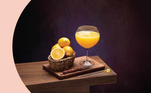 A shot of a superjuice in a coupe glass on a wooden board and table surrounded by knife and four oranges in a basket