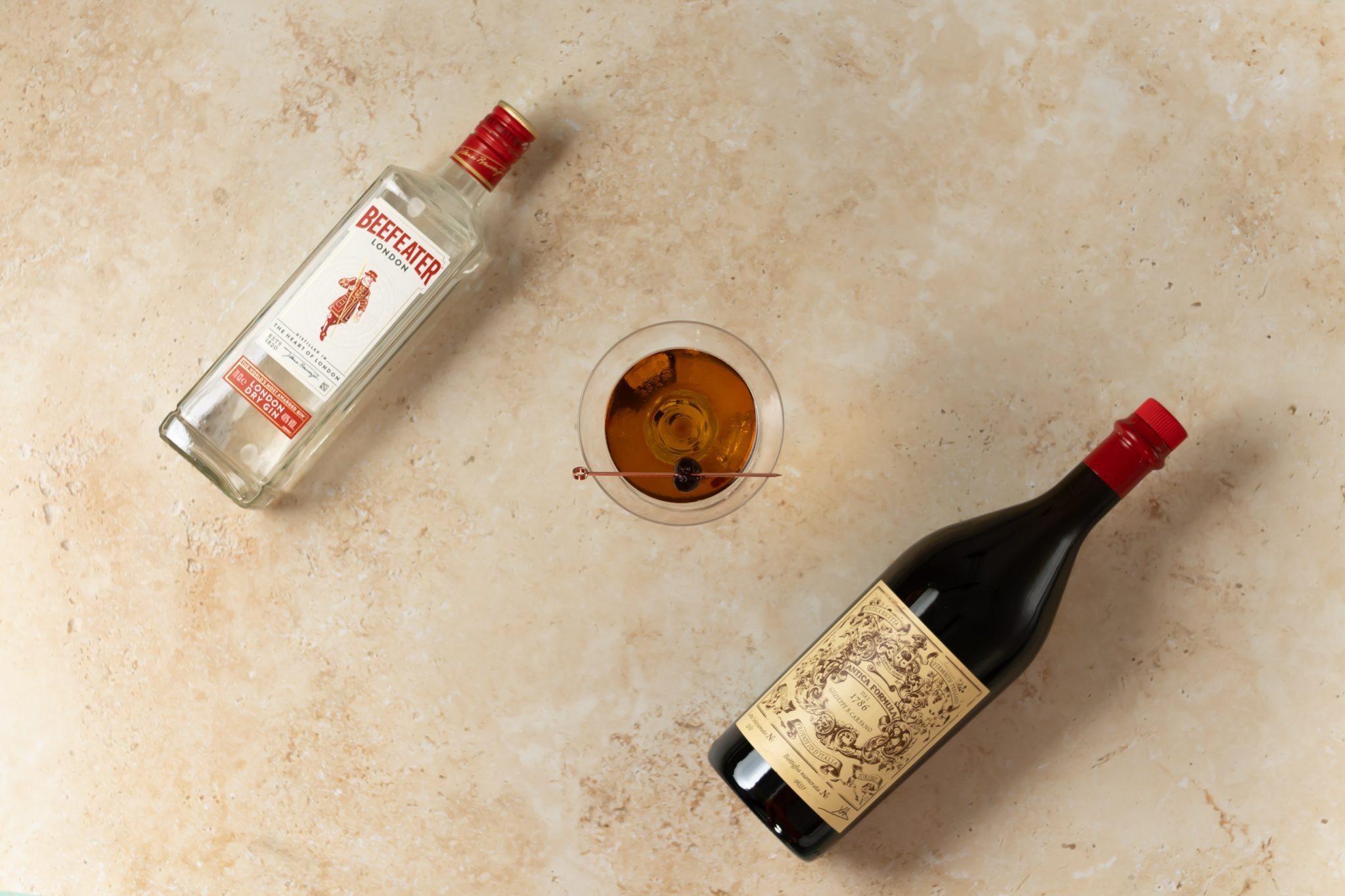 A Sweet Martini cocktail, shot from above, in a martini glass on a beige surface surrounded by Gin and sweet Vermouth