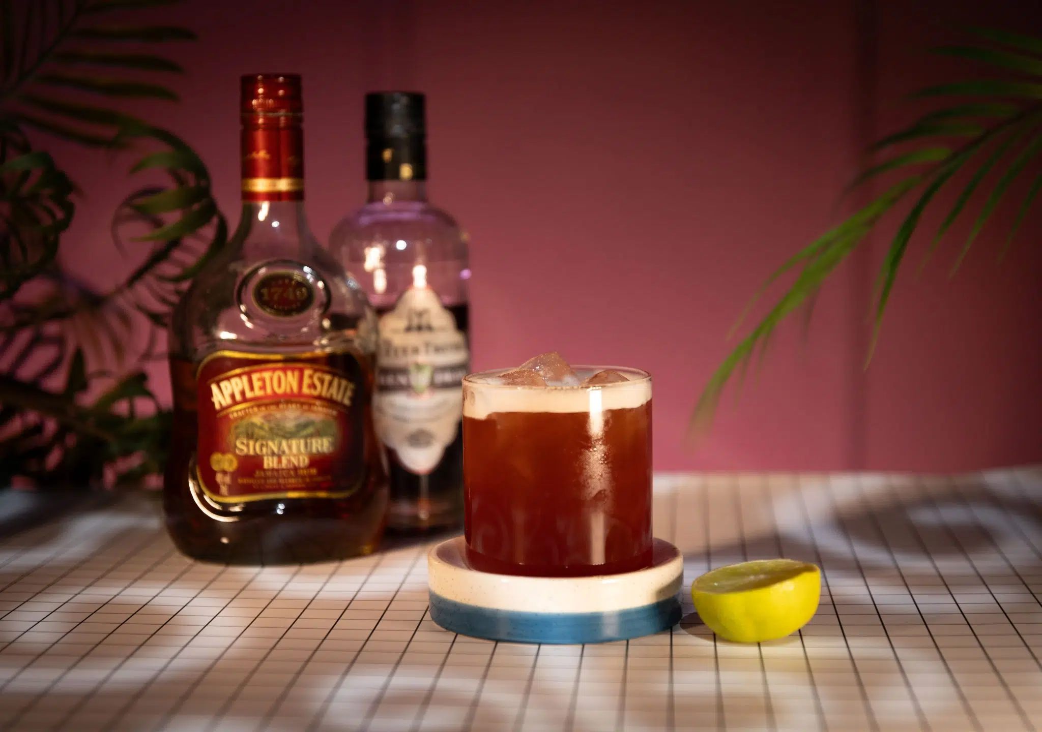 A side shot of a Tea and Krampus cocktail in a rocks glass on a blue and white coaster surrounded by aged dark rum bottle, allspice dram liqueur bottle and half a lemon