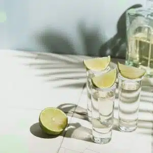 Three tall shot glasses with salt on the rim and a lime wedge on top filled with tequila on a white surface with a transparent bottle of tequila in front of a light blue wall.