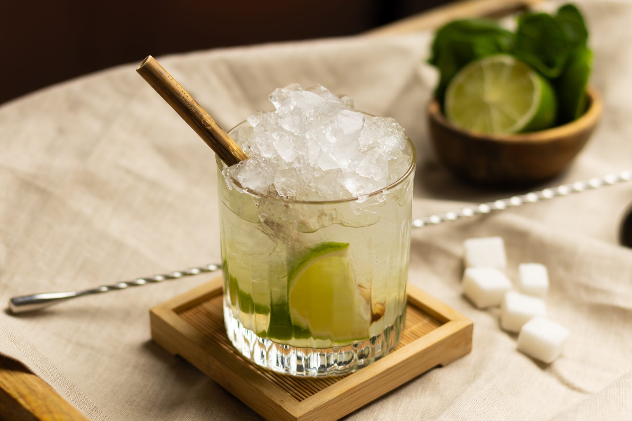 A side shot of a Tequila Caipirinha cocktail in an old fashioned glass on a wooden coaster surrounded by a withe cloth a bar spoon, a bowl with lime pieces, and five sugar cubes