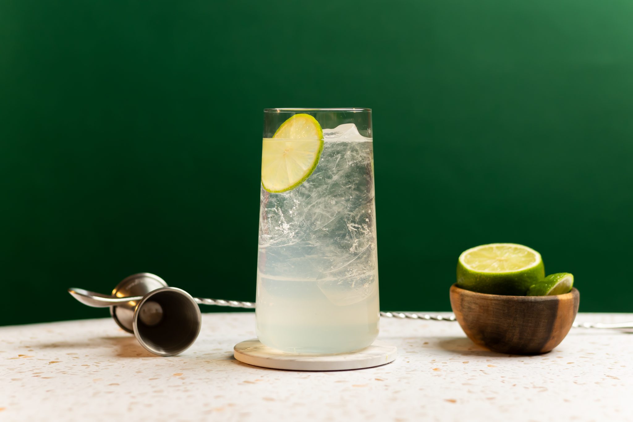 A side shot of a Tequila Cooler cocktail in a highball glass on a coaster placed on a white table surrounded bya jigger, a bar spoon, and a bowl with limes, in front of a green background