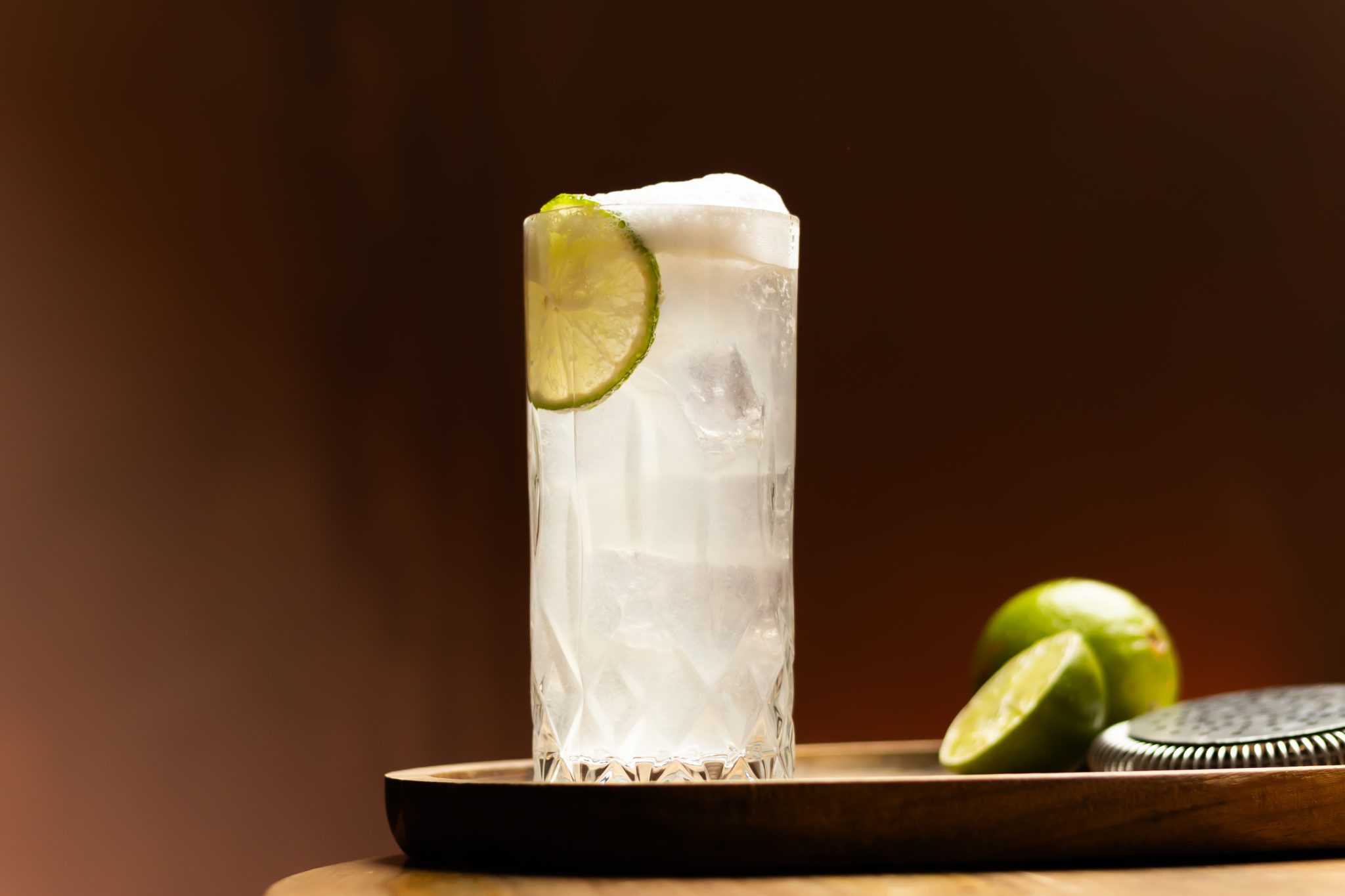 A side shot of a Tequila Fizz cocktail in a highball glass on a wooden board with two limes and a cocktail strainer on the side