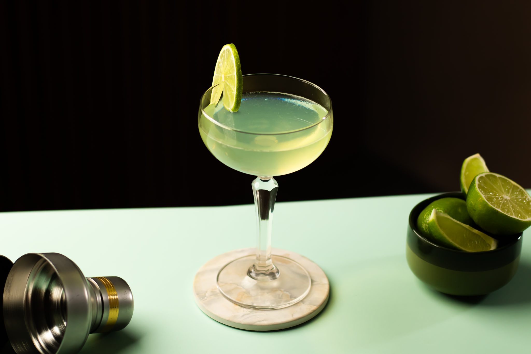A side shot of a Tequila Gimlet cocktail in a cocktail glass on a white coaster placed on a light green table surrounded by a bowl with limes and a shaker.