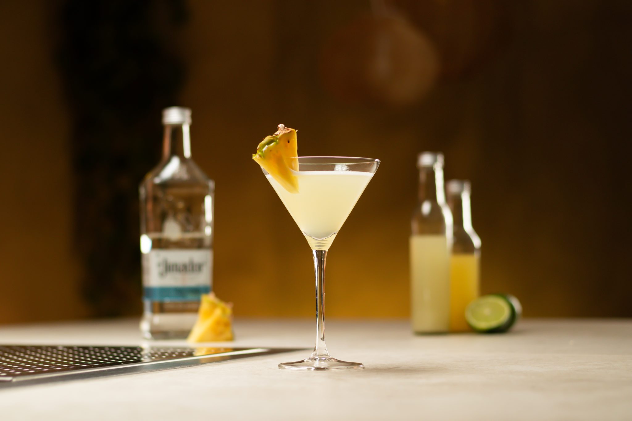 Tequila, pineapple juice, lime juice, two limes and two pineapple pieces laid out on a white bar table