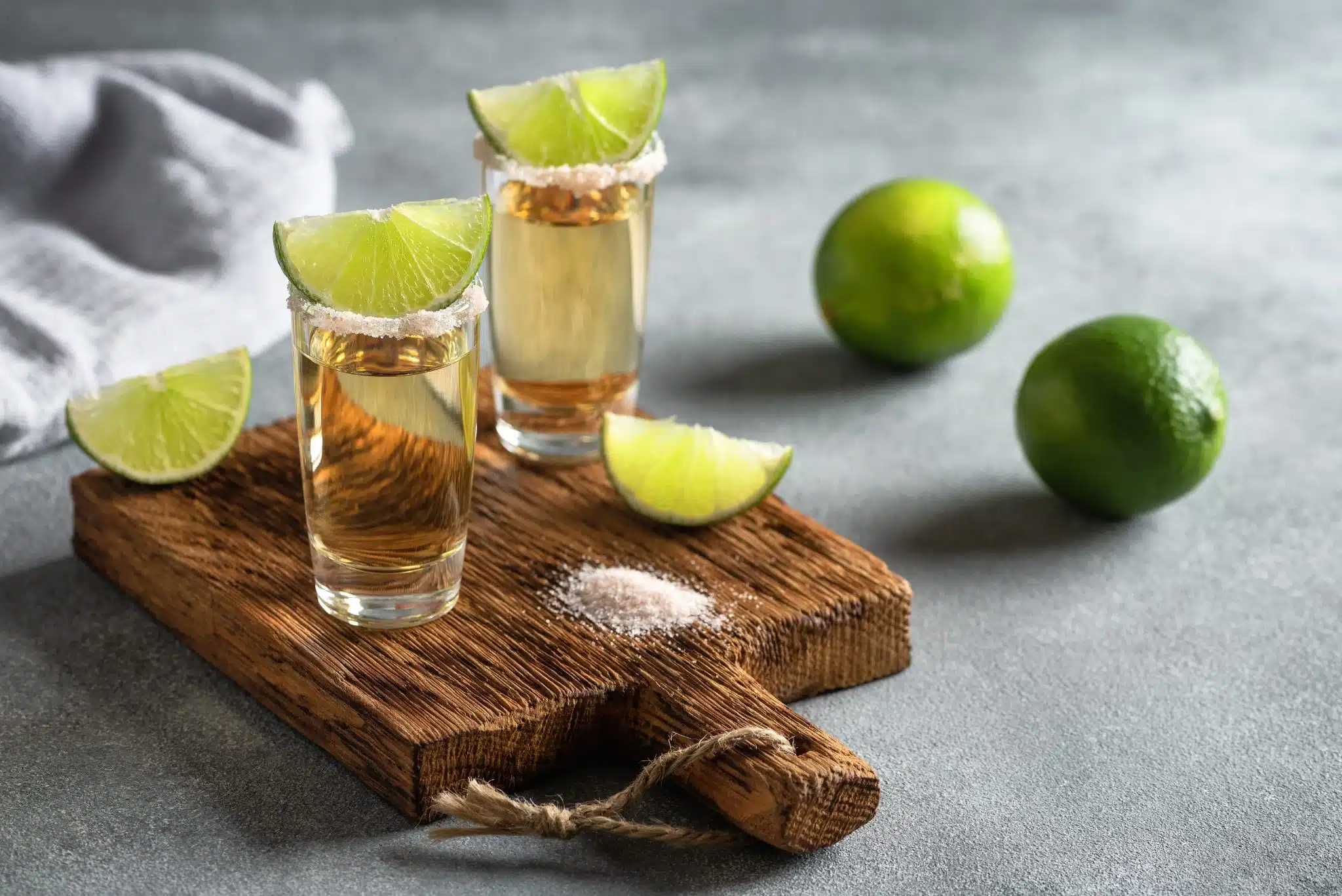 Two shots of tequila on a wooden board with lime wedges and salt.