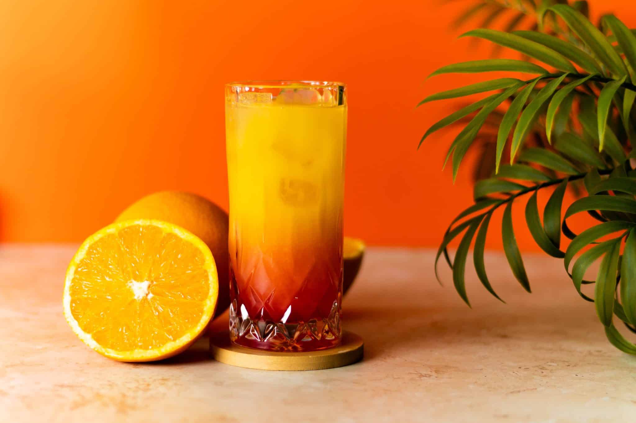 A side shot of a Tequila Sunrise cocktail in a highball glass on a brown coaster placed on a beige table with a half orange on one side and a green plant on the other side.