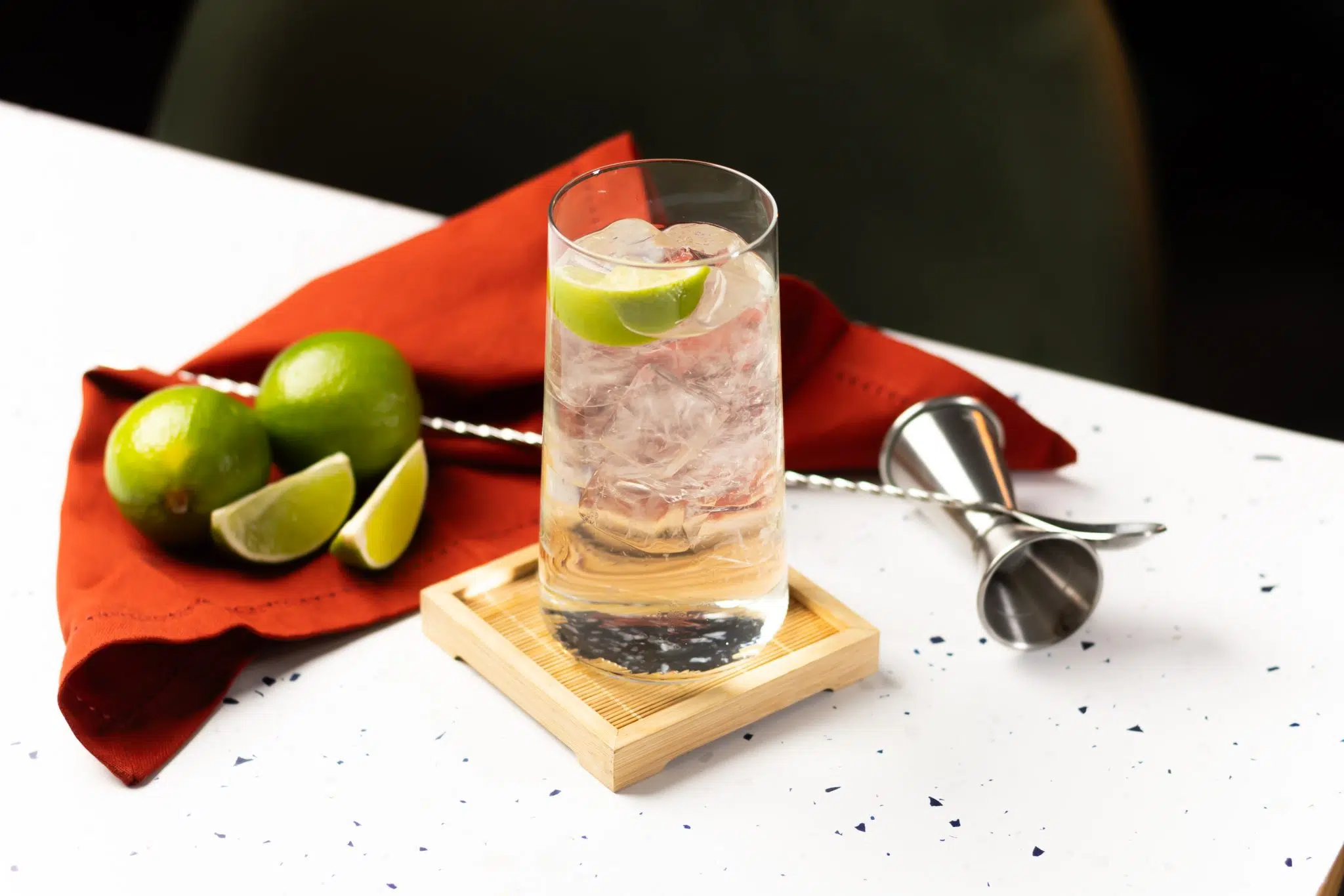 A side shot of a Tequila Tonic cocktail in a highball glass on a wooden coaster placed on a white bar table surrounded by a jigger, a bar spoon, two limes, two lime wedges anda red cloth.