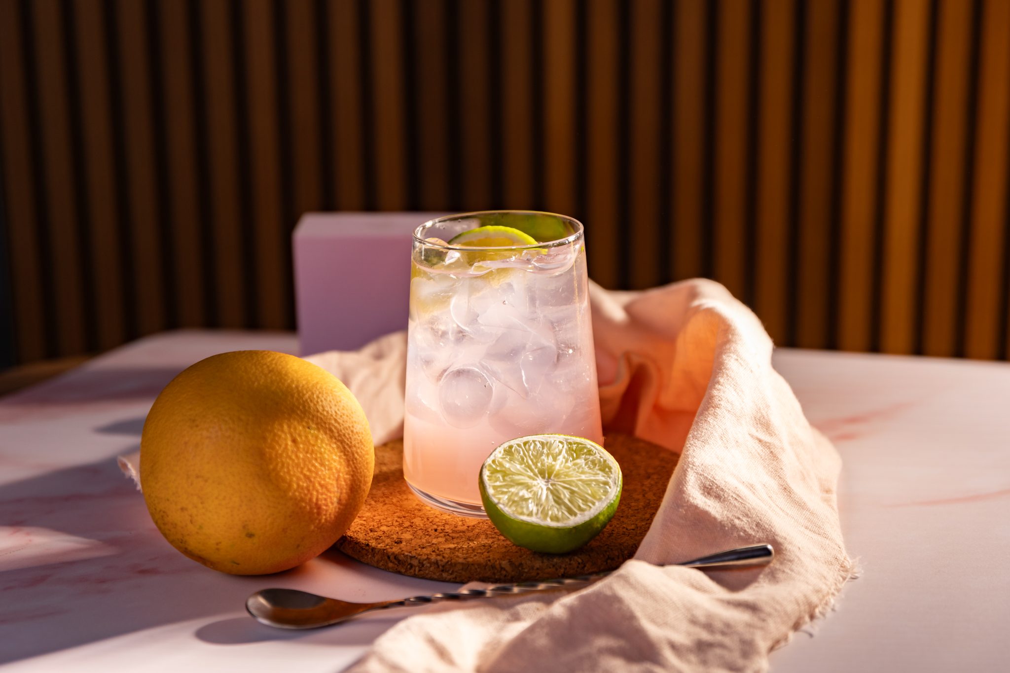 A side shot of a Tequila and Grapefruit Soda cocktail in a highball glass on a cork coaster placed on a white surface surrounded by half lime, an orange, a bar spoon and a pale pink cloth