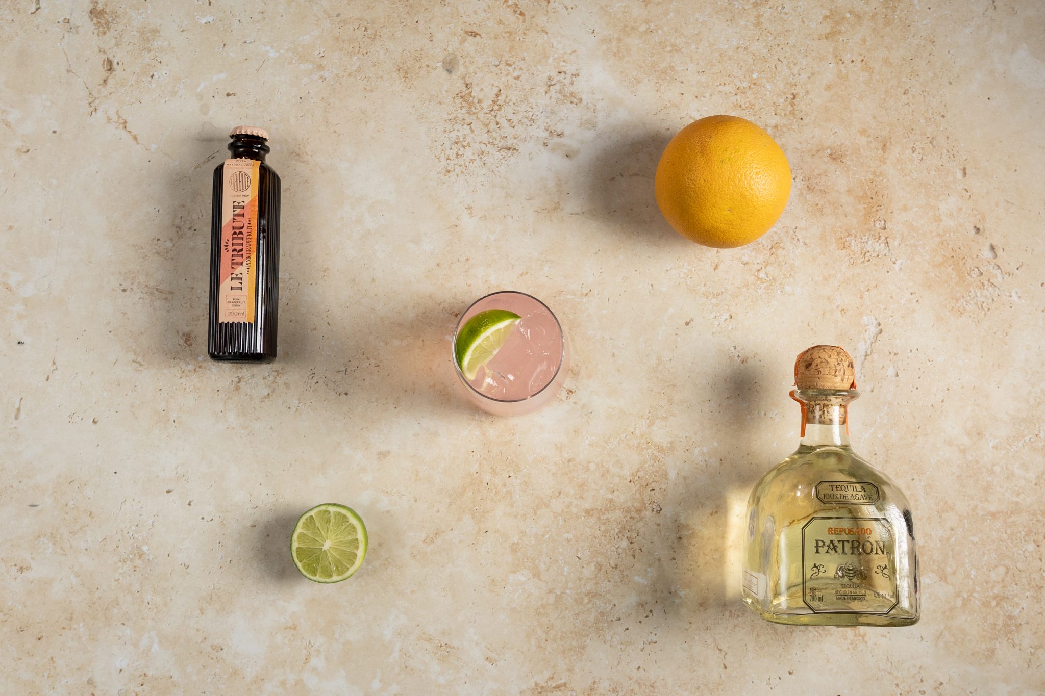 A Tequila and Grapefruit Soda cocktail, shot from above, in a highball glass on a beige surface surrounded by tequila, grapefruit soda, lime and orange