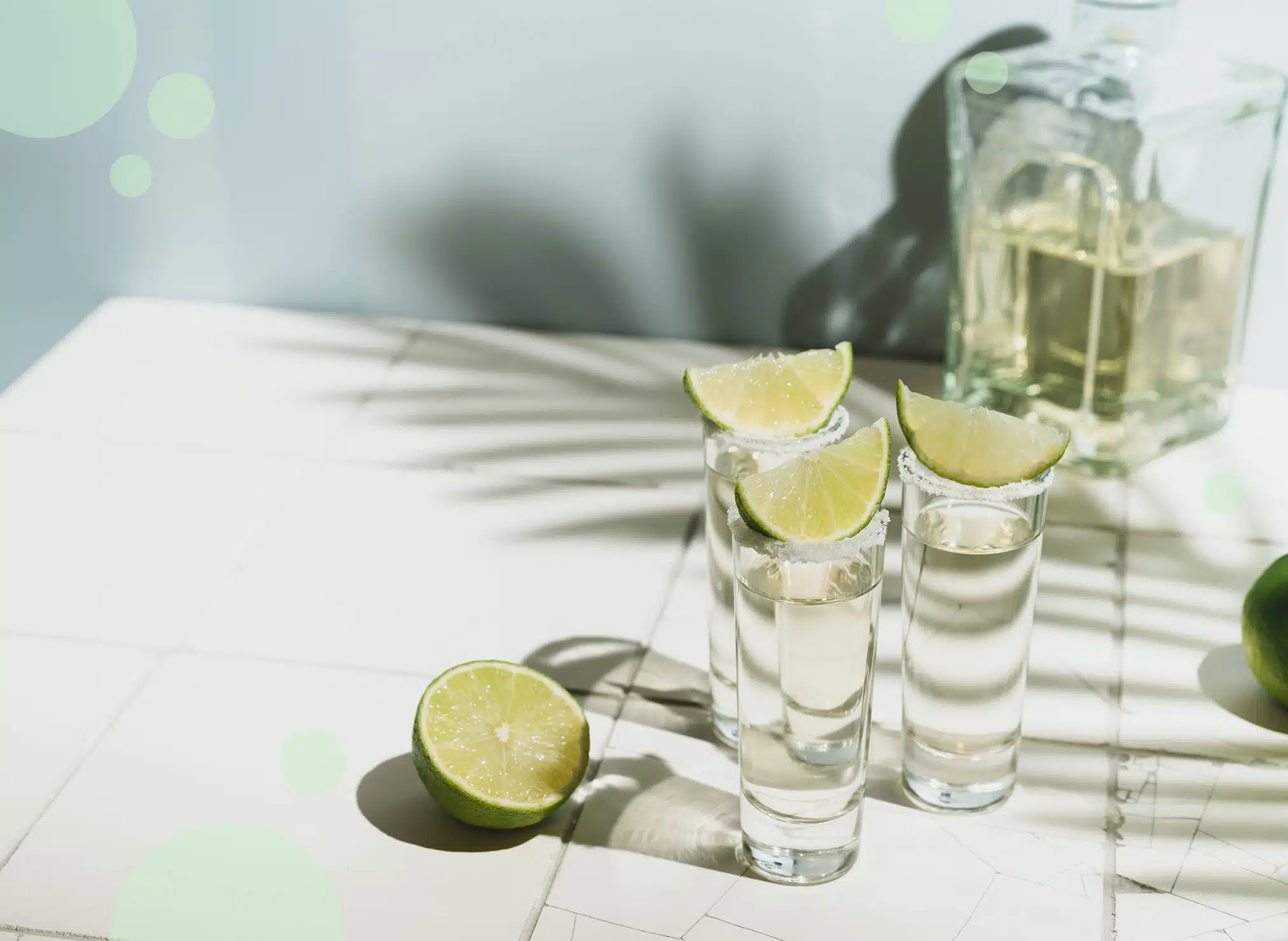 Three tall shot glasses with salt on the rim and a lime wedge on top filled with tequila on a white surface with a transparent bottle of tequila in front of a light blue wall.