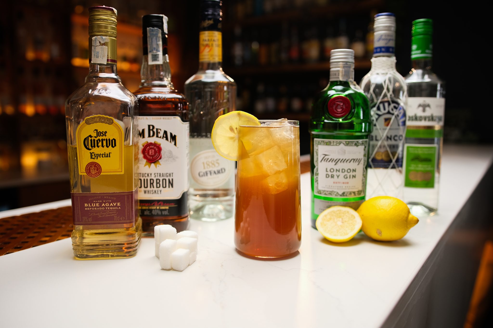 Tequila, Bourbon, Triple Sec, Gin, Rum, Vodka, sugar cubes and two lemons laid out on a white bar table.