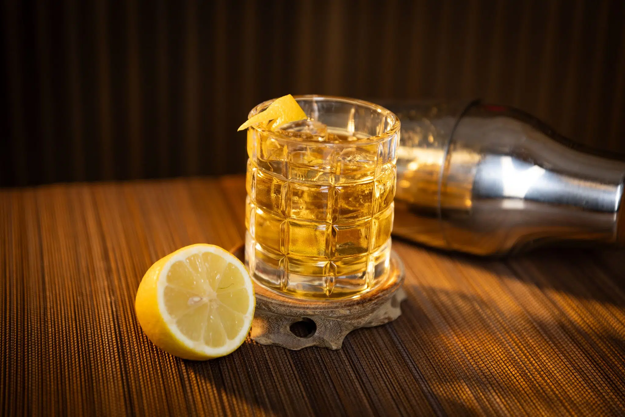 A side shot of The Beast cocktail in an old fashioned glass on a wooden coaster and surface surrounded by a cocktail shaker and half a lemon