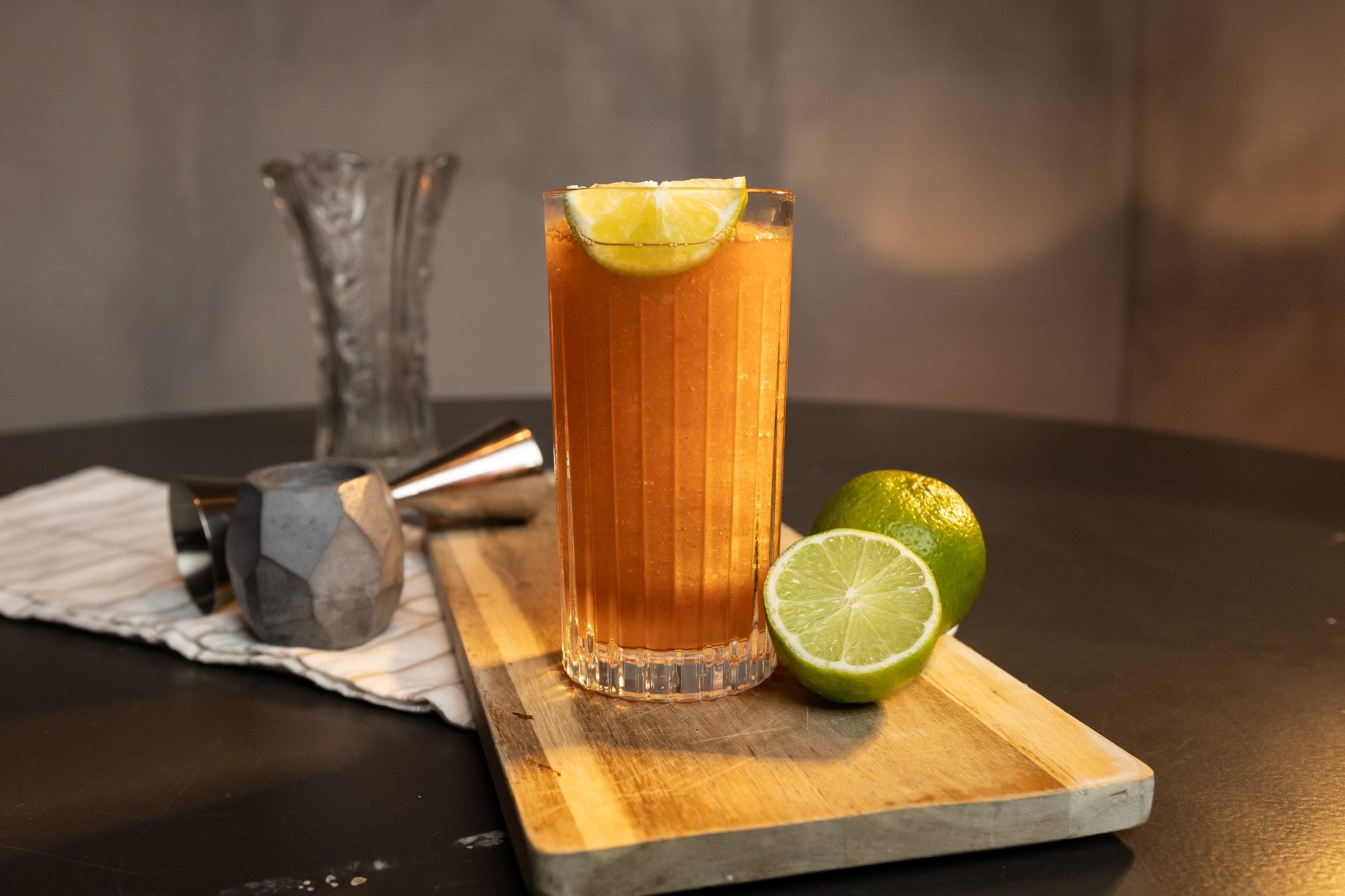 A side shot of a Tobago Tea cocktail in a highball glass on a wooden board surrounded by two limes, a jigger, a glass, a pot and a white cloth