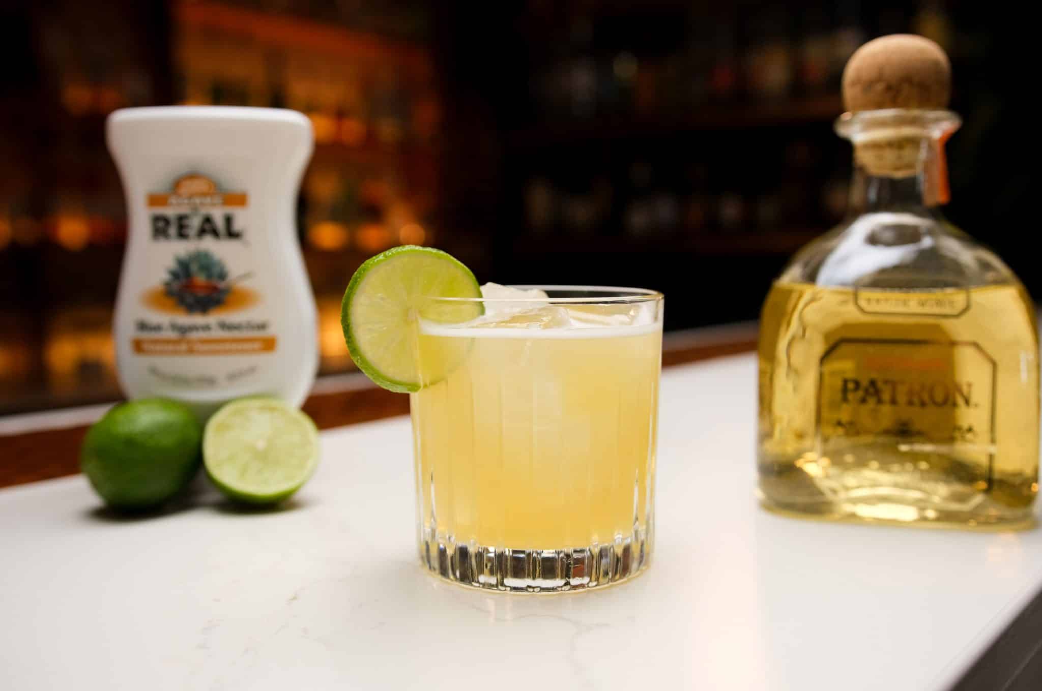 Tequila, agave nectar and two limes laid out on a white bar table.