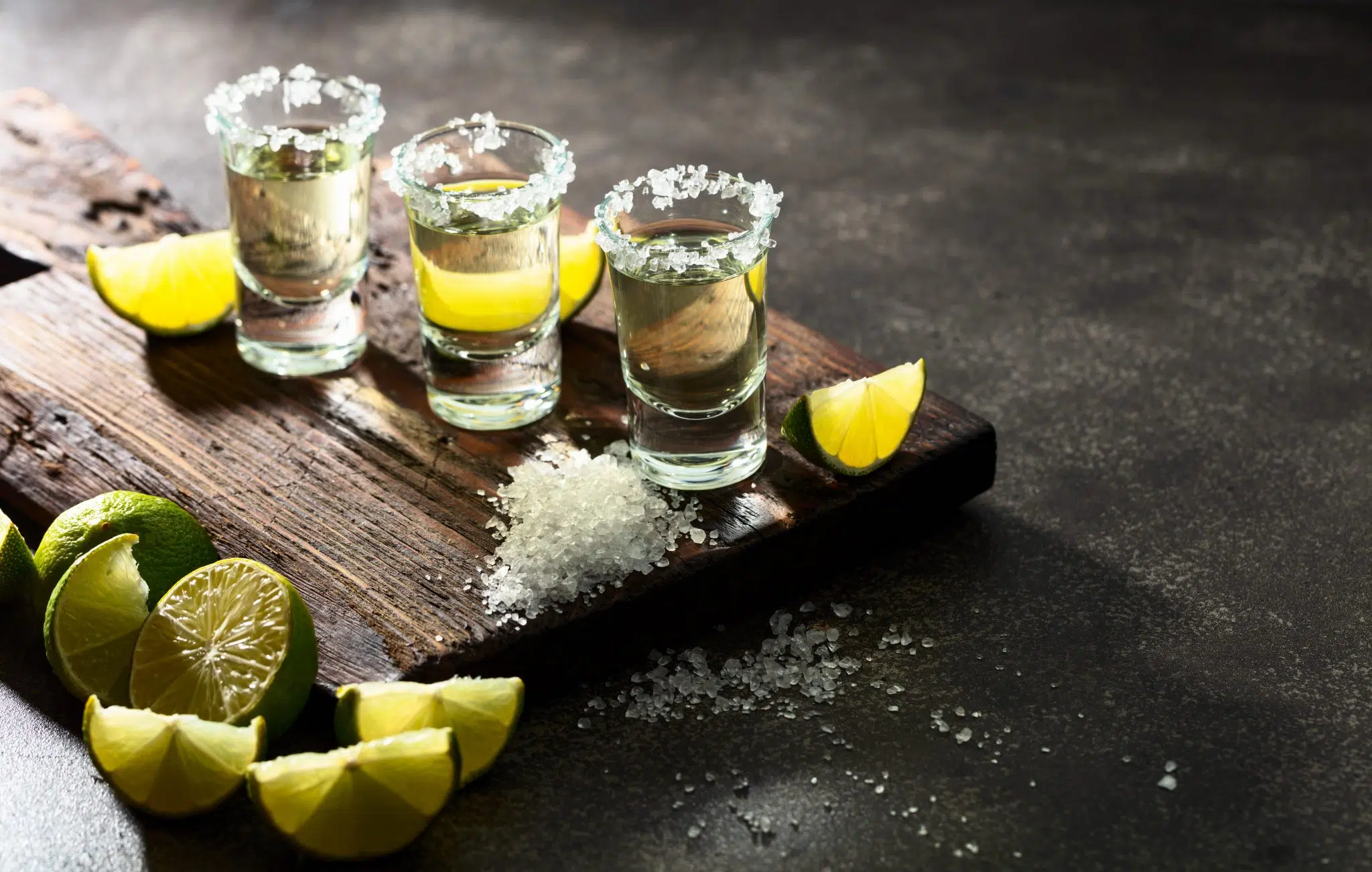 A side shot of three traditional tequila shots with lime wedges and salt around, on top of a wooden board and a dark grey surface
