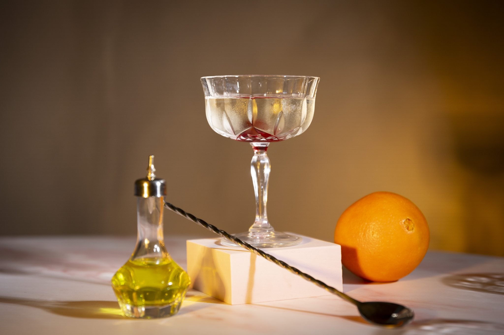 A side shot of a Tuxedo No.2 cocktail in a coupe glass on a white tall and square coaster placed on a white surface surrounded by a bar spoon, and absinthe bottle and an orange