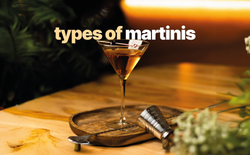 A side shot of a martini cocktail in a martini glass on a wooden board and table surrounded by a jigger and a bar spoon