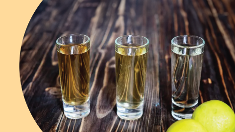 A side shot of three shot glasses with different types of tequila on a wooden surface and two lemons on the side