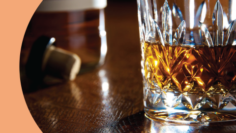 A shot of whiskey cocktail in an Old Fashioned glass on a dark wooden surface with a cork and a bottle on the side