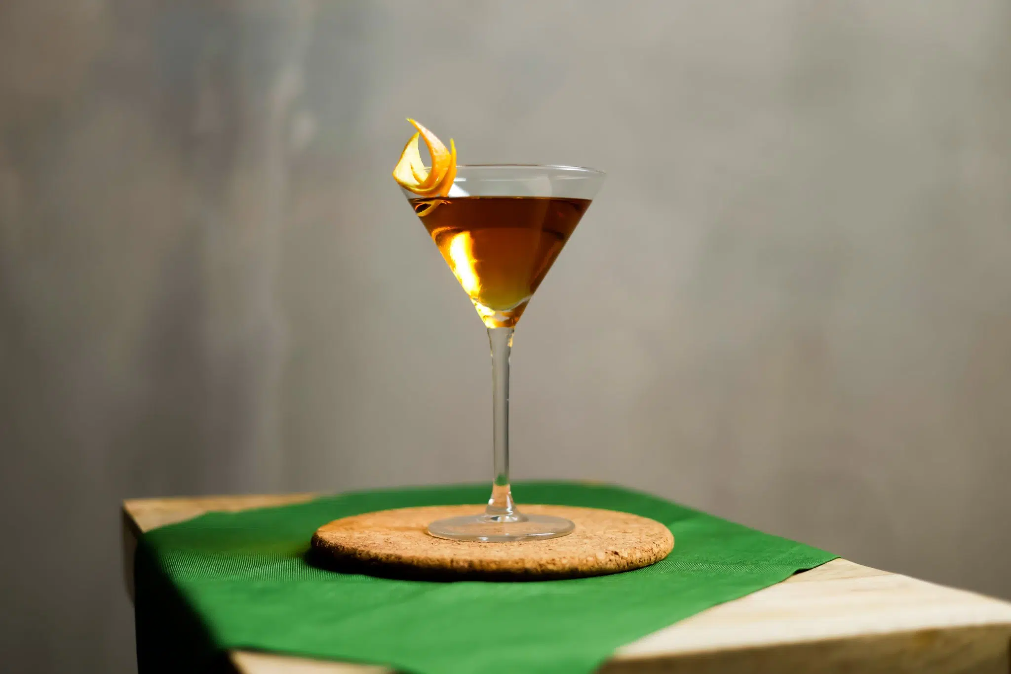 A side shot of a Vancouver cocktail in a martini glass on a cork coaster placed over a green placemat in front of a grey background