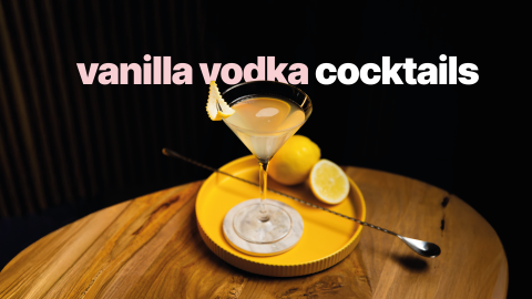 A shot of a vanilla vodka cocktail in a martini glass on a white coaster on a yellow tray placed on a wooden table in front of a black background
