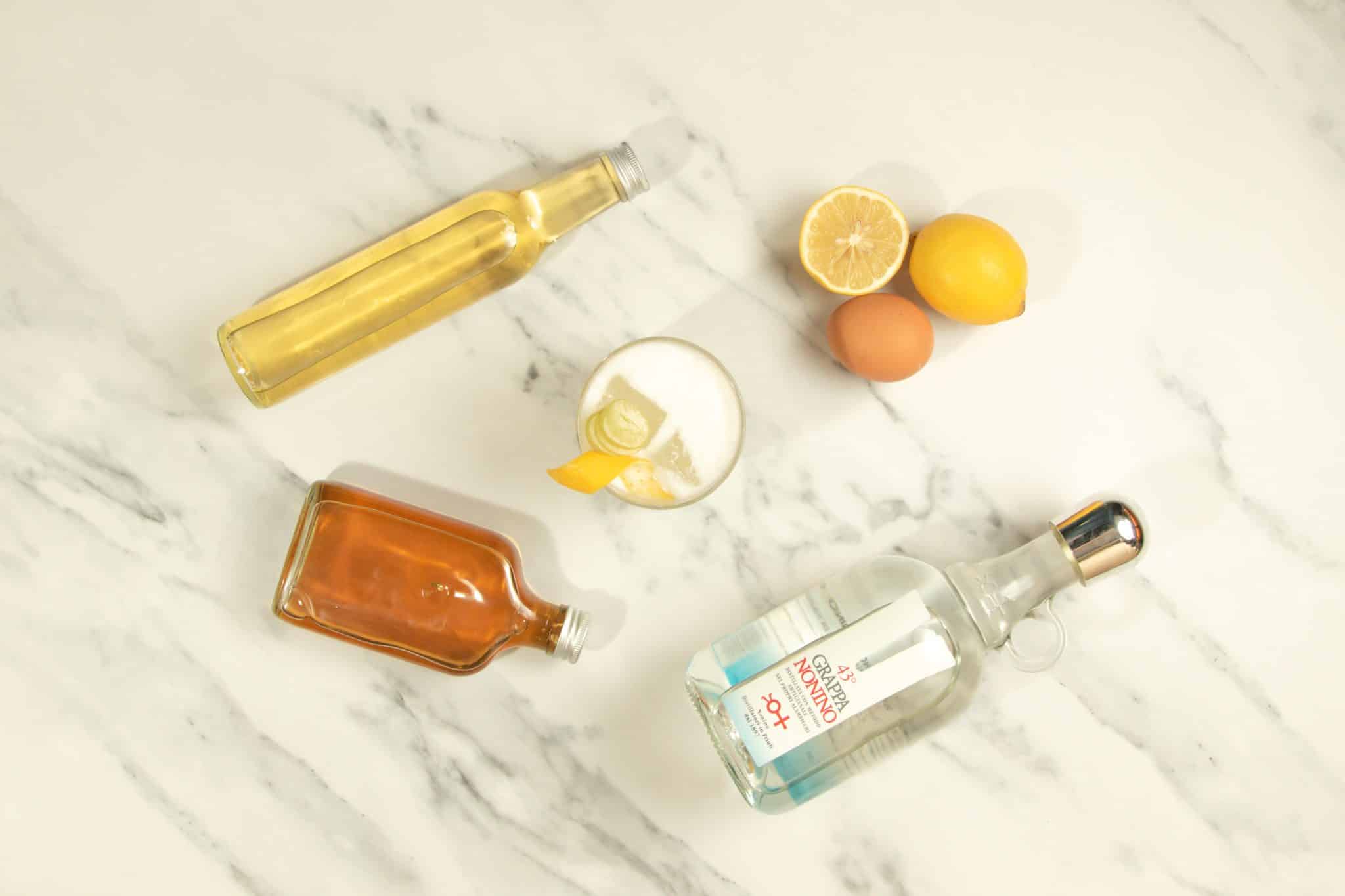 A VE.N.TO cocktail on a white marmol table with a White Grappa bottle, a honey mix bottle, a chamomile cordial bottle, an egg and two lemons around.