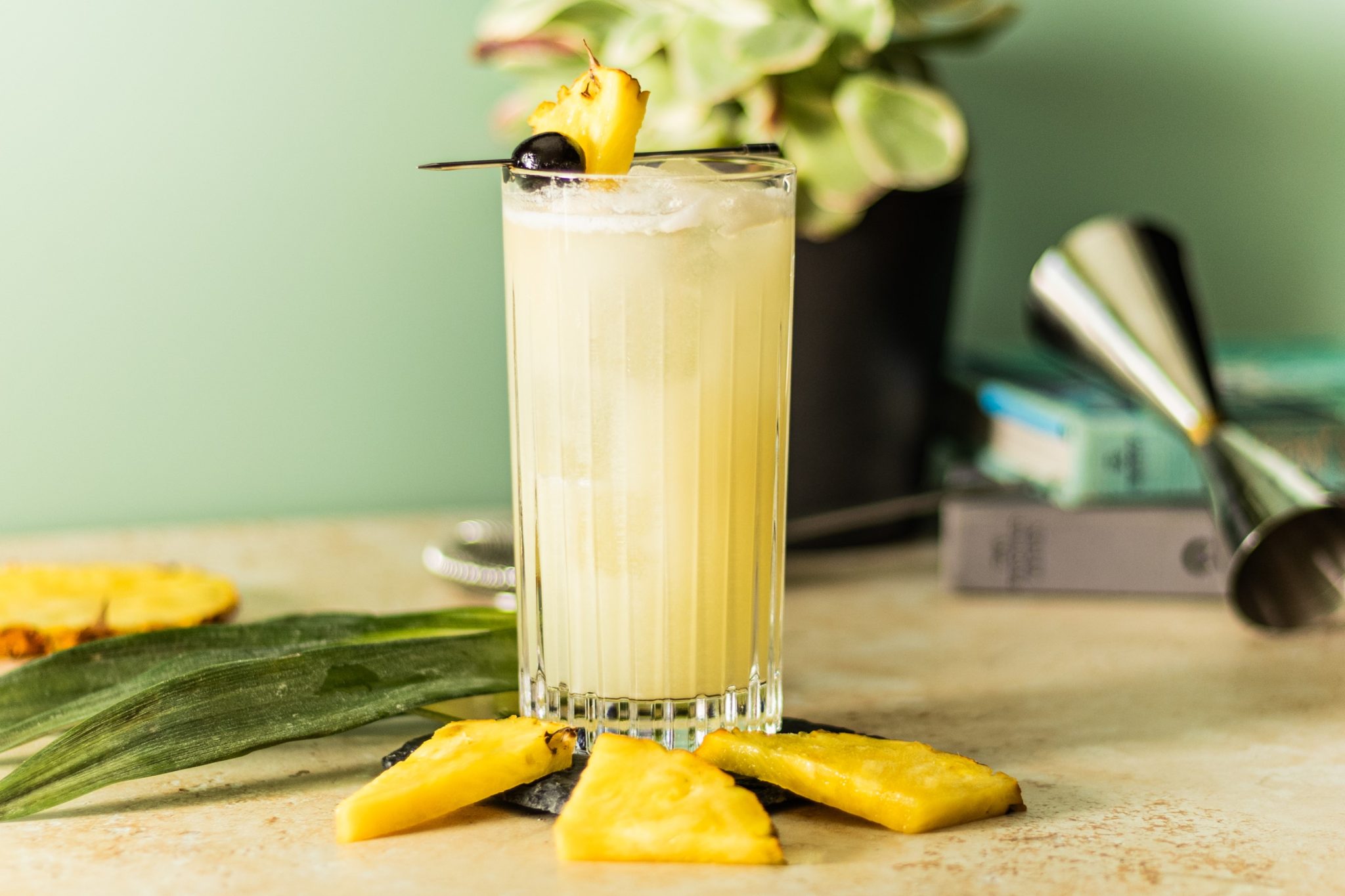 A side shot of Virgin Piña Colada Cocktail in highball glass on a beige table surrounded by three pineapple slices, some plant leaves, two books and a jigger, in front of a light green wall.