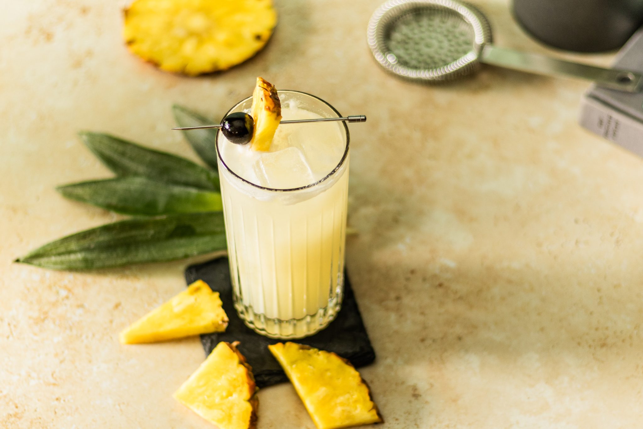A Virgin Piña Colada Cocktail in highball glass on a black stone coaster placed on a beige table surrounded by three pineapple slices, a pineapple wheel, some plant leaves, and a strainer.