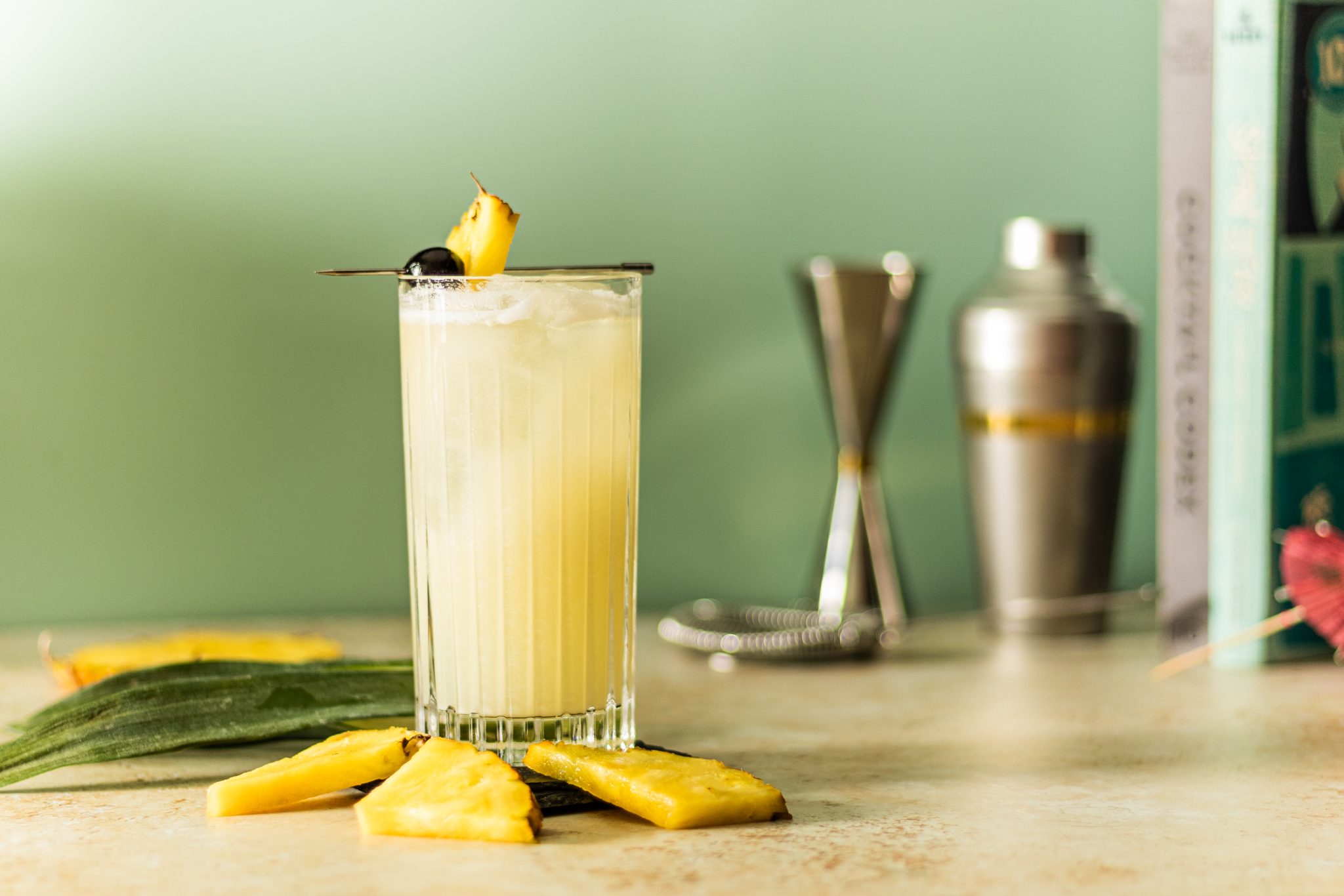 A side shot of Virgin Piña Colada Cocktail in highball glass on a beige table surrounded by three pineapple slices, some plant leaves, two books, a shaker, and a jigger, in front of a light green wall.