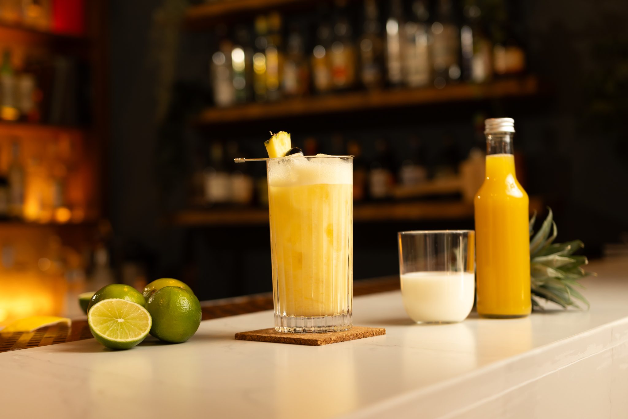 Pineapple juice, coconut cream, and two limes on a white bar table.