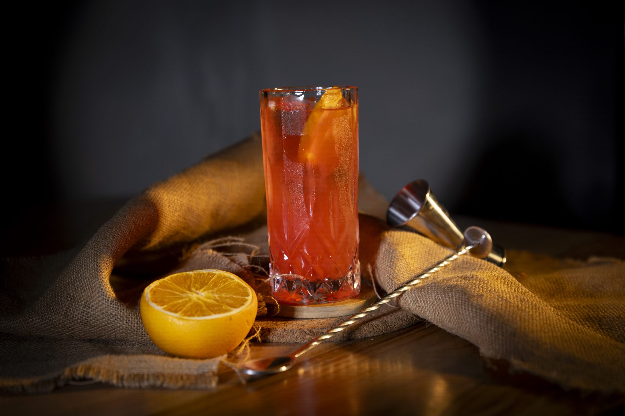 A side shot of a Vodka Cran-Orange cocktail in a highball glass on a wooden coaster placed on a wooden surface surrounded by half orange, a bar spoon, a jigger and a brown cloth