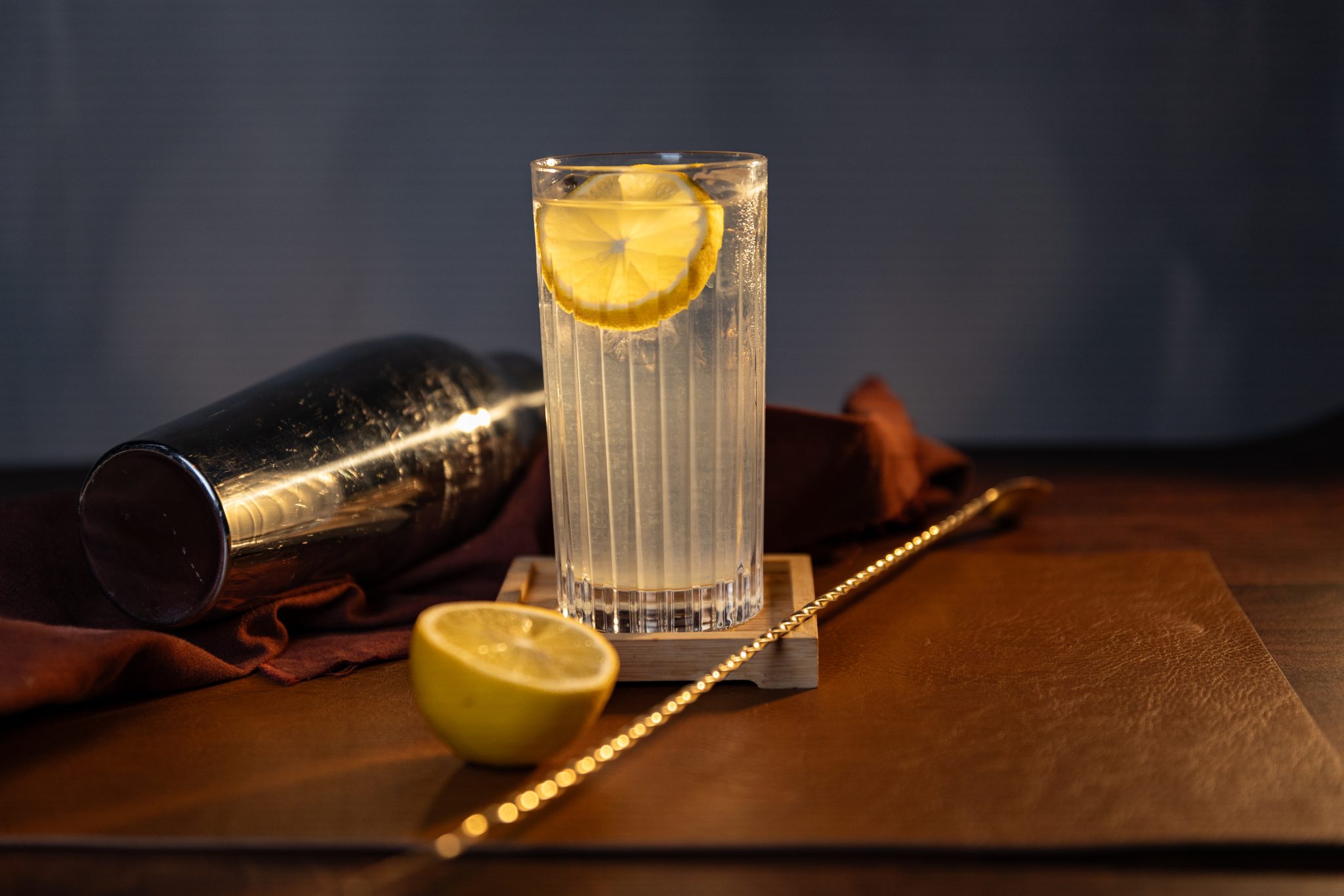 A side shot of a Vodka Lemonade cocktail in a highball glass on a wooden surface surrounded by a bar spoon, a cocktail shaker, a brown cloth and half lemon