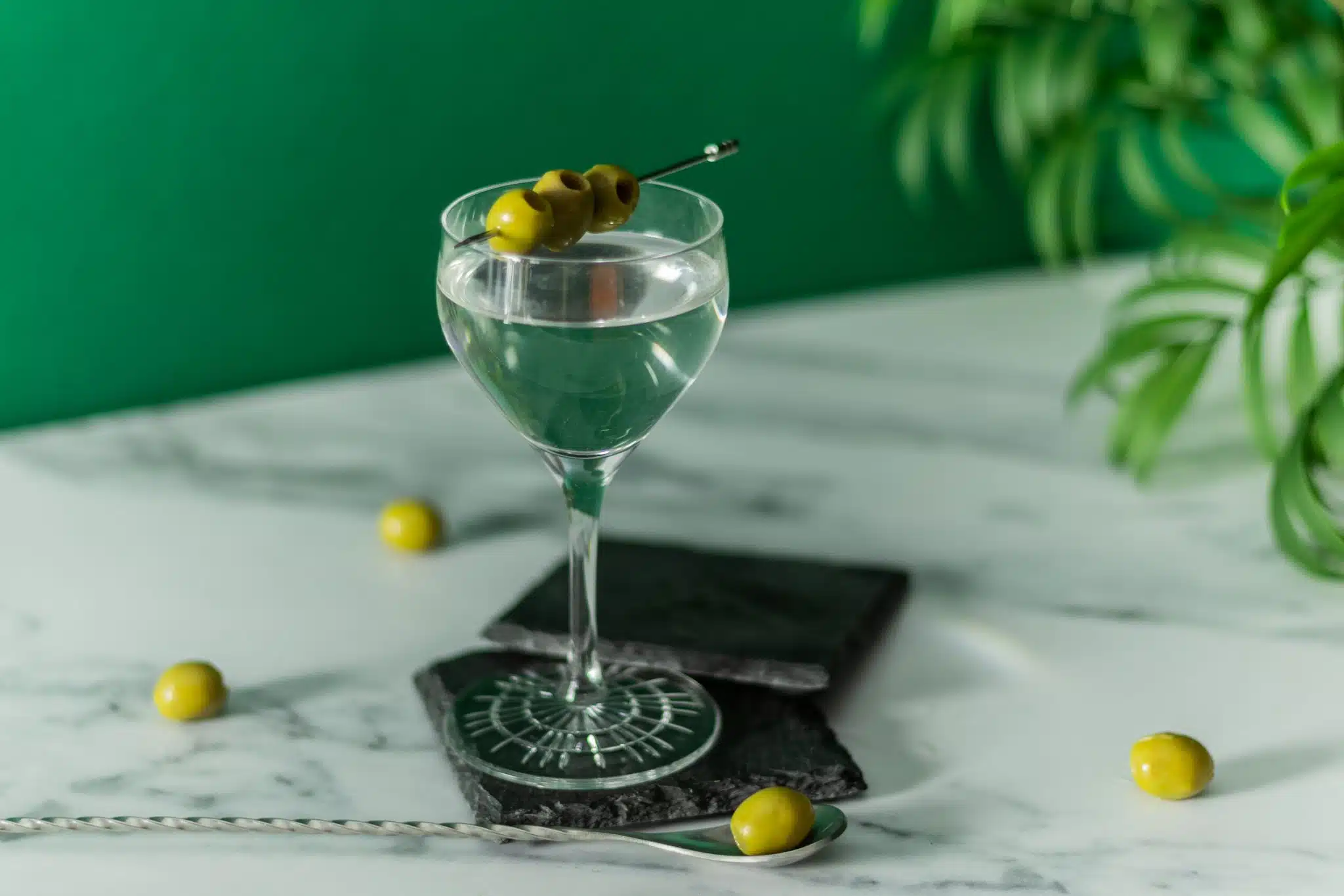 A side shot of a Vodka Martini cocktail garnished with three olives on a stone plate placed on a marmol table 