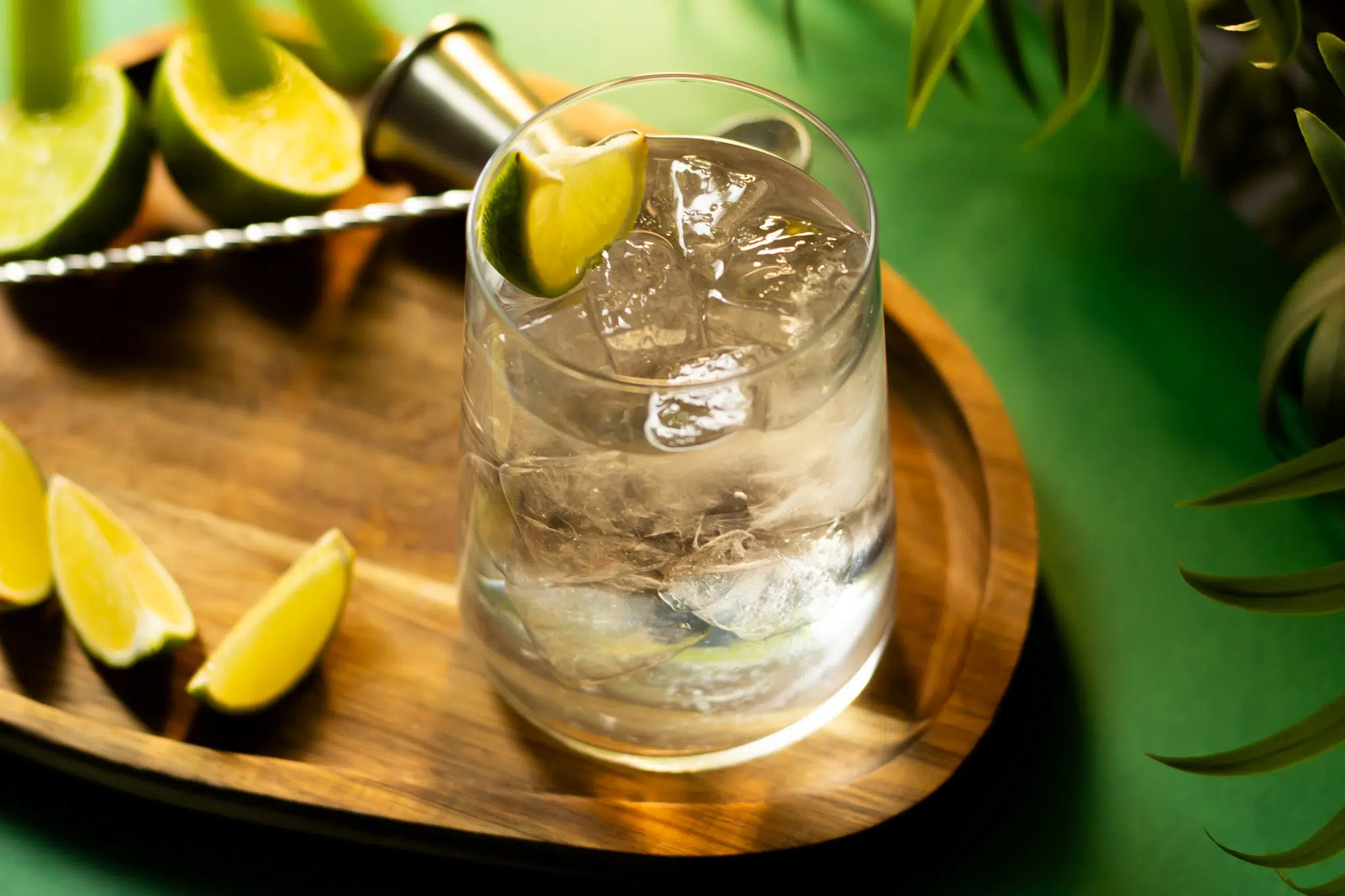 A high angle shot of a Vodka Soda cocktail in highball glass on a wooden board placed on a green surface surrounded by three lime wedges, two half limes, a jigger and a bar spoon