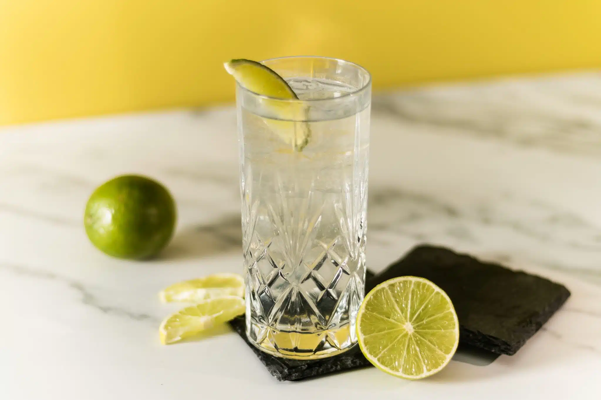 A side shot of a Vodka Tonic cocktail in highball glass on a black stone coaster with lime pieces around, placed on a white marmol table in front of a yellow wall.