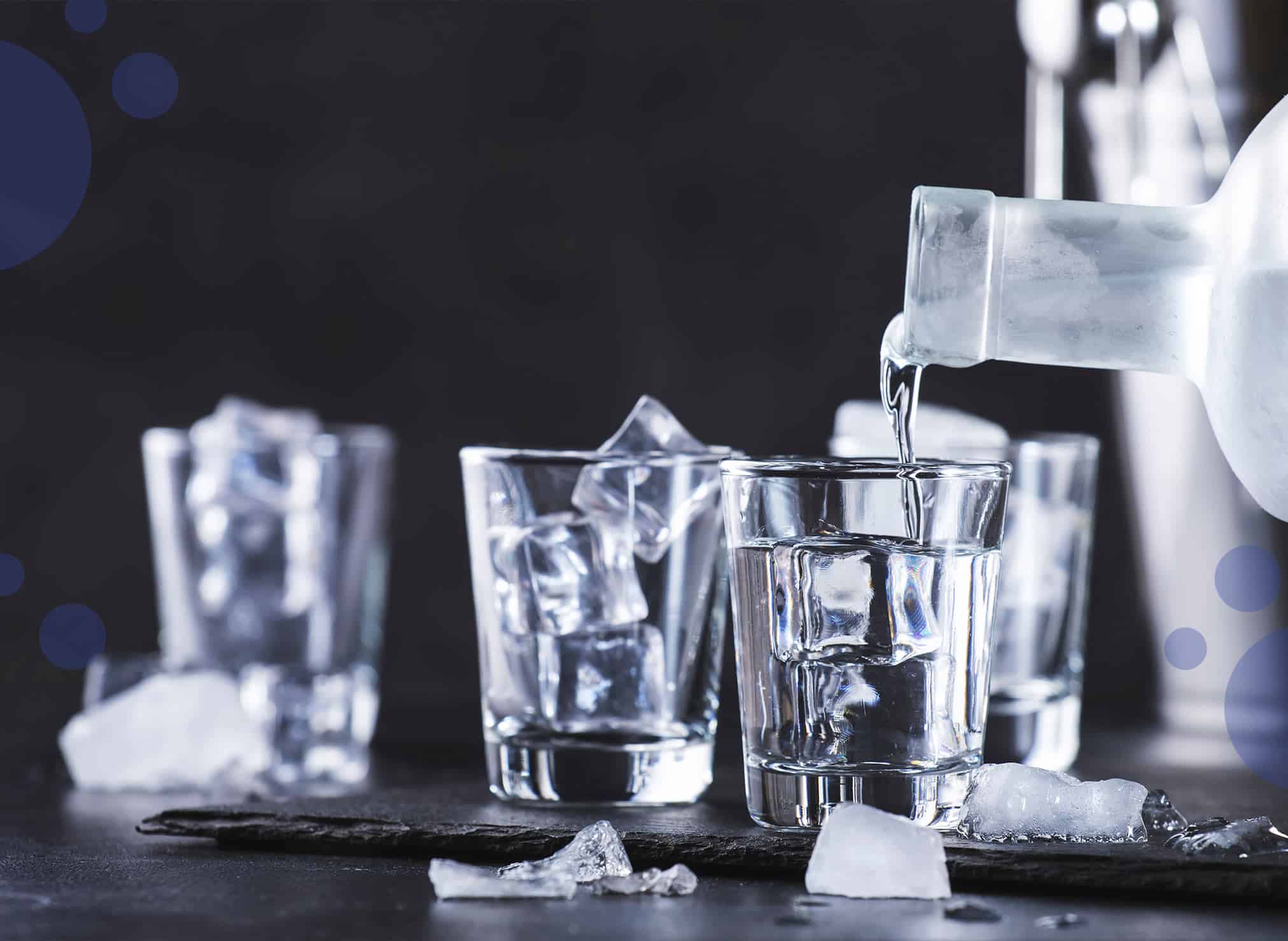4 shot glasses being filled with ice cubes and chilled vodka on a black stone plate placed on a black table in front of a black background.