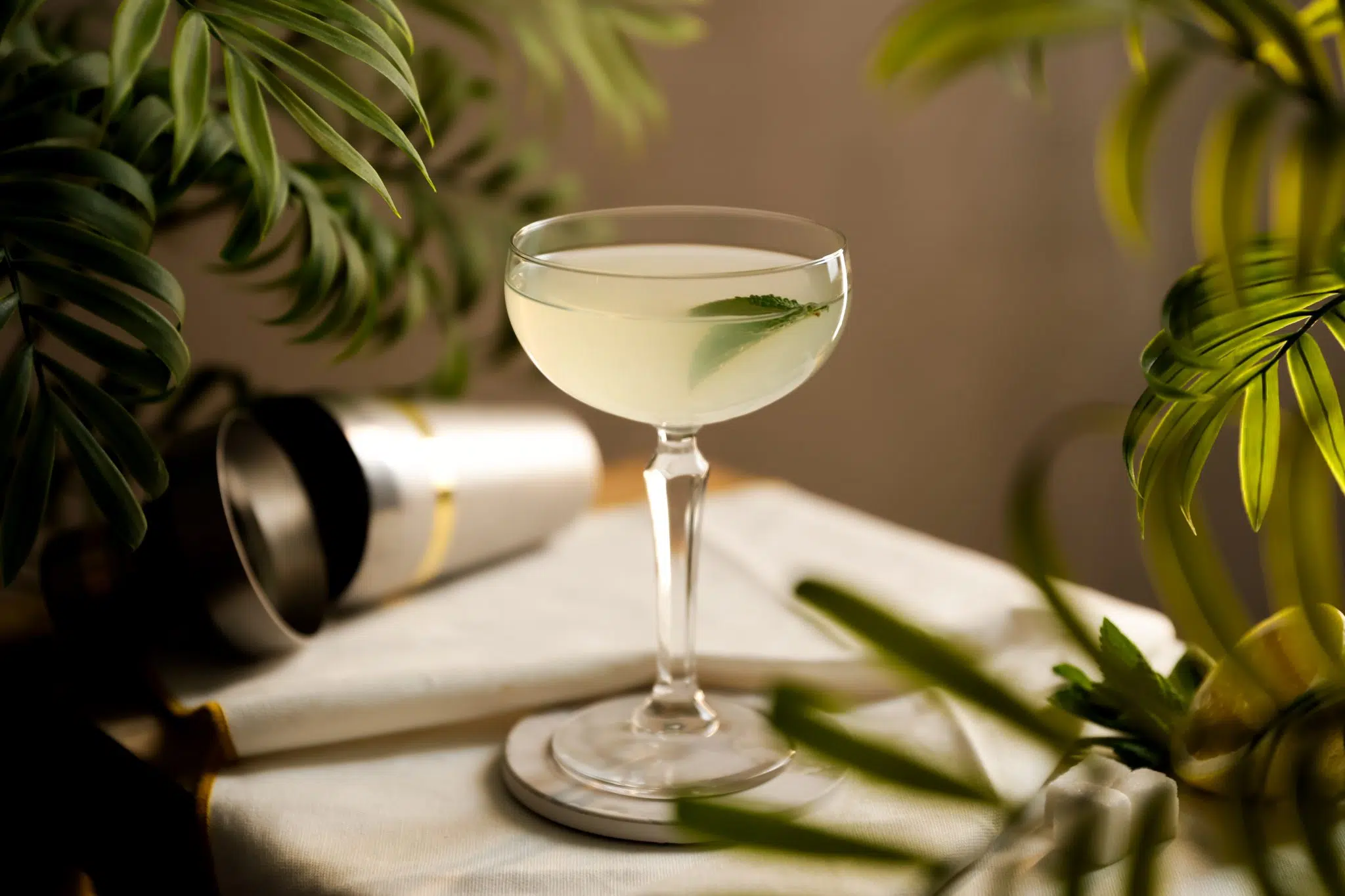 A side shot of a West Side cocktail in a coupe glass on a white coaster with a white cloth and a cocktail shaker behind
