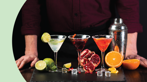 A shot of three Tequila cocktails in martini glasses on a black stone board surrounded by two limes, mint leaves, half pomegranate, two half oranges, four ice cubes and a cocktail shaker, and in front of a person with a red shirt and black pants