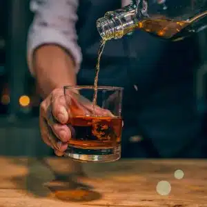Old-fashioned glass holded by a hand, being filled with whiskey from a transparent bottle over a wooden bar table.