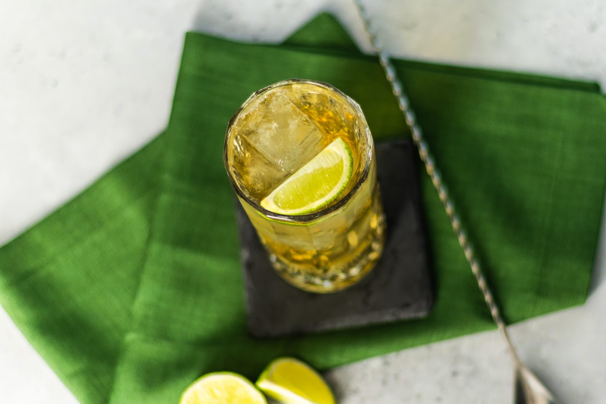 A Whiskey Ginger cocktail, shot from above, in a highball glass on a black stone coaster placed on a green cloth on a white table with two lime wedges and a bar spoon around.