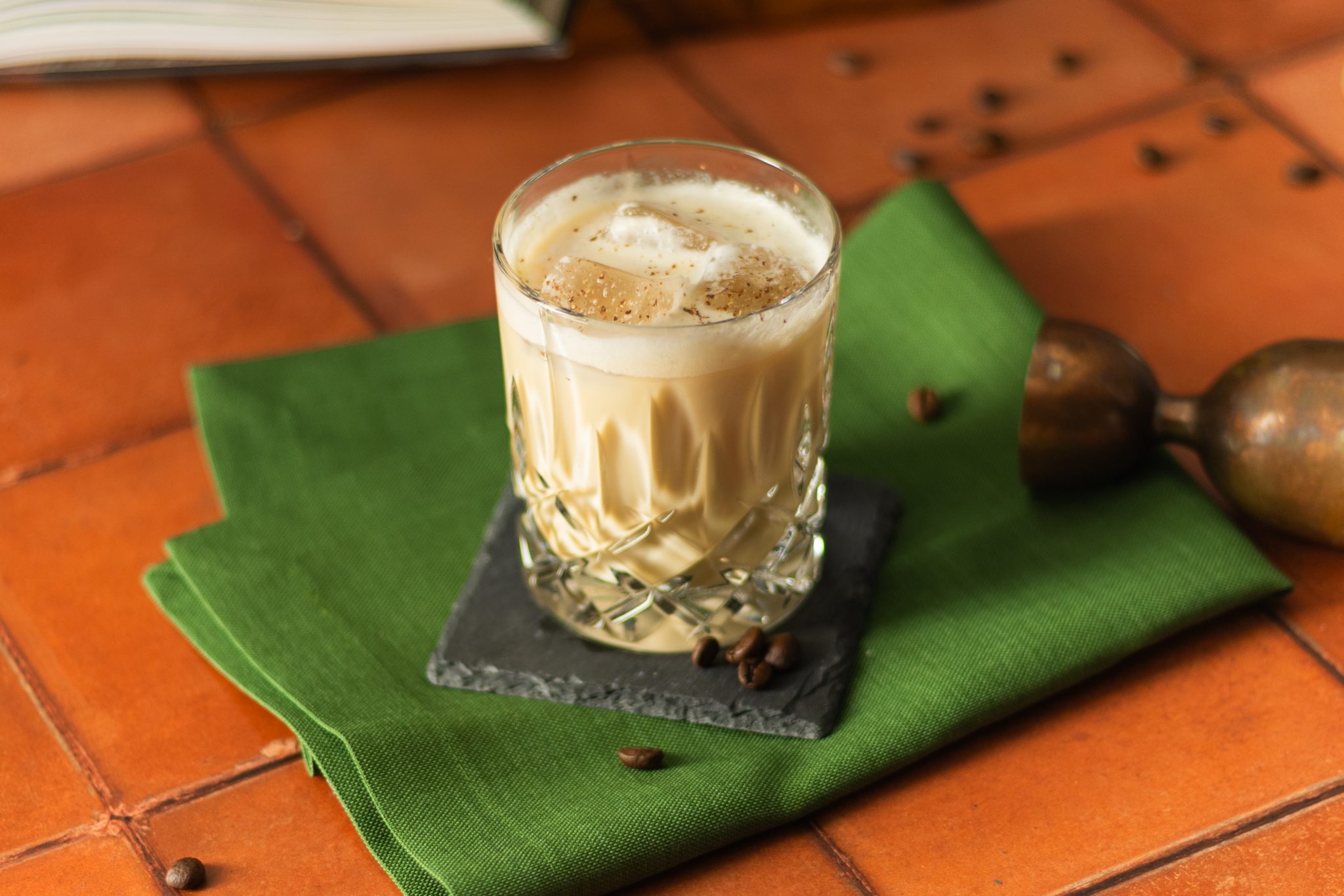 A White Cuban cocktail, shot from above, in an old fashioned glass on a green cloth placed on a tiled surface, surrounded by coffee beans, and a jigger.