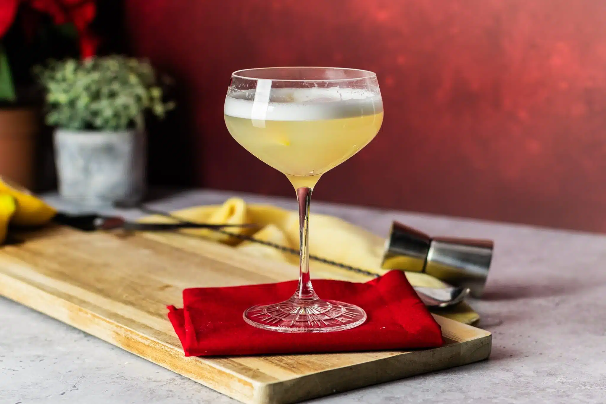A side shot of a White Lady cocktail in a coupe glass on a red cloth placed on a wooden tray on top of a white table with a jigger, a bar spoon and a yellow cloth around.