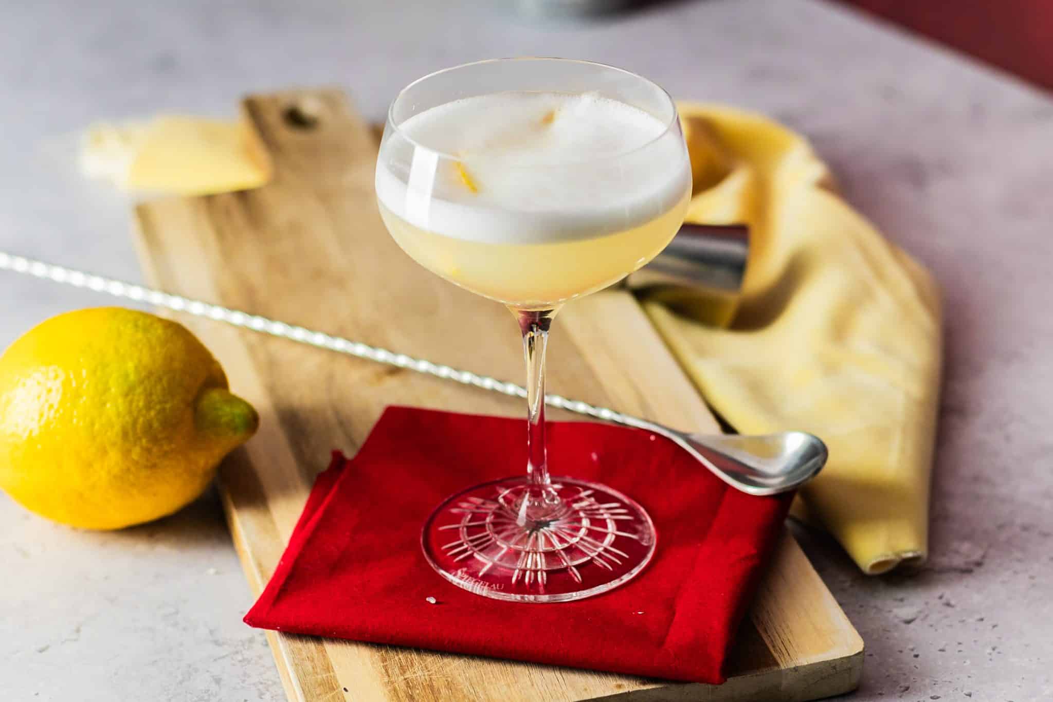 A side shot of a White Lady cocktail in a coupe glass on a red cloth placed on a wooden tray on top of a white table with a jigger, a bar spoon, a lemon and a yellow cloth around.