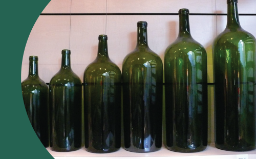 A side shot of six green wine bottles in different sizes in front of a wall